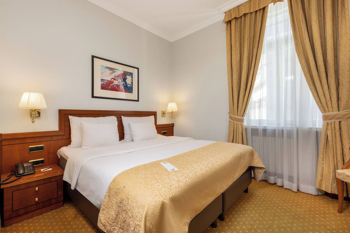 Ein Hotelzimmer mit einem Bett, Nachttischen, Lampen, einem gerahmten Bild an der Wand und einem Fenster mit Vorhängen.