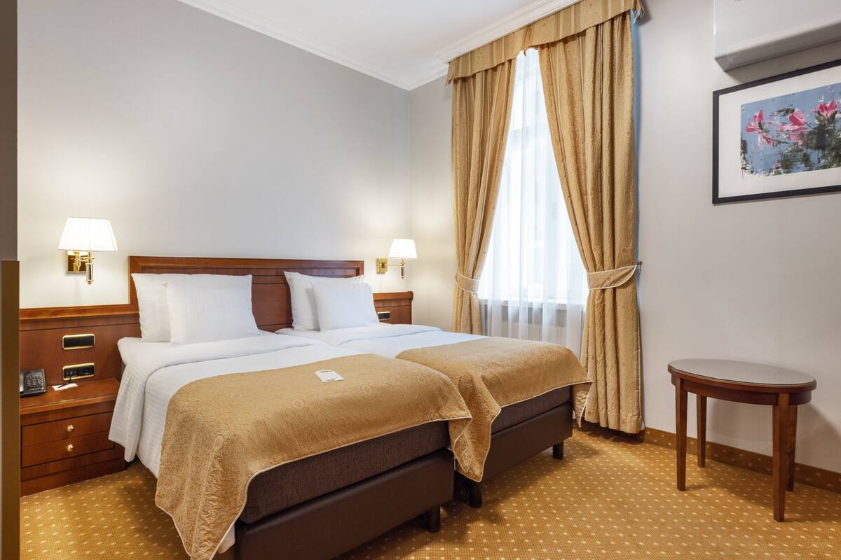 Ein Hotelzimmer mit Doppelbett, zwei Nachttischen, einem kleinen runden Tisch und einem großen Fenster mit Vorhängen.