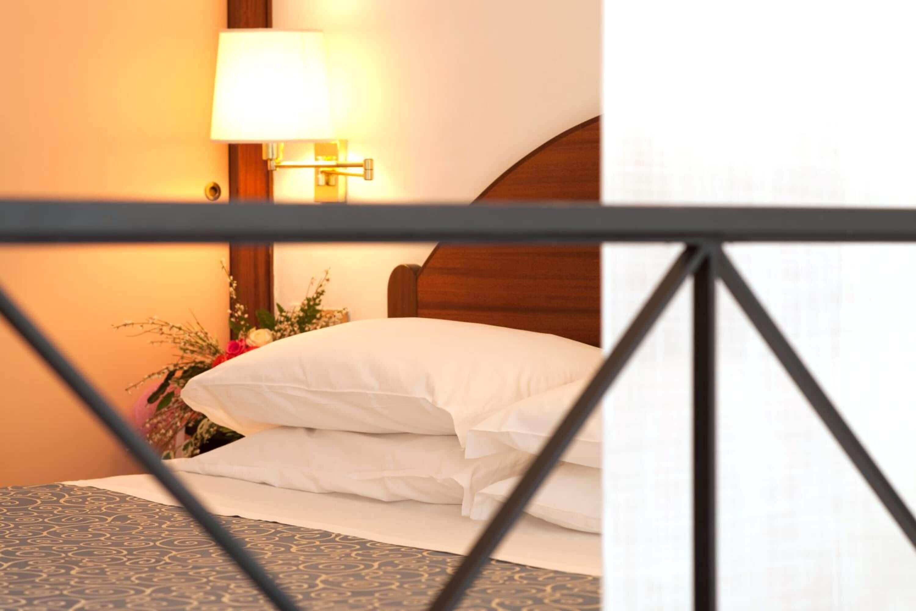 A hotel room with a neatly made bed, a lamp on the nightstand, and a floral arrangement.