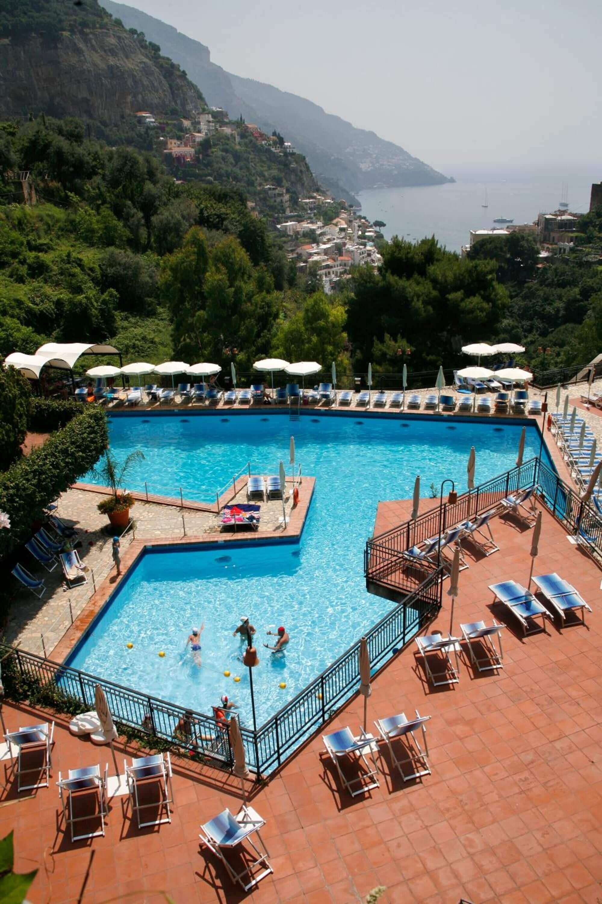 A swimming pool with multiple lanes, surrounded by sun loungers and umbrellas, set against a backdrop of a coastal town and hills.