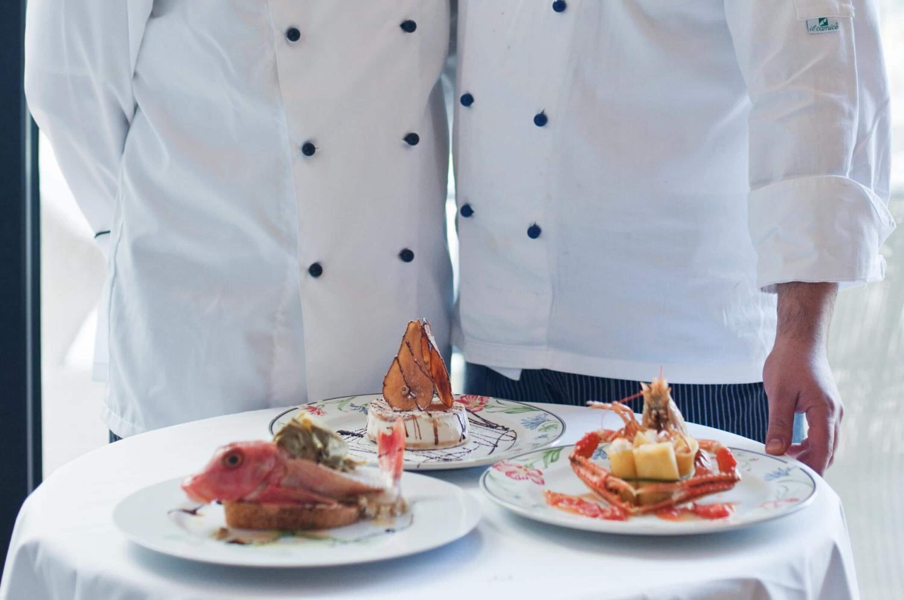 Un chef con uniforme blanco y botones negros de pie detrás de una mesa con platos de mariscos.