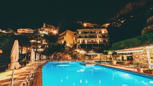 A swimming pool with lounge chairs and umbrellas, surrounded by buildings with balconies and lights, indicating a resort setting. - Hotel Royal Positano (Positano)