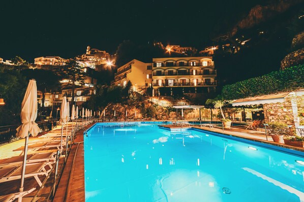 A swimming pool with lounge chairs and umbrellas, surrounded by buildings with balconies and lights, indicating a resort setting.