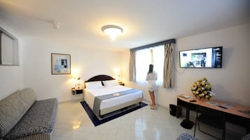 Habitación de hotel con cama, sofá, escritorio con computadora, televisor y ventana con cortinas.