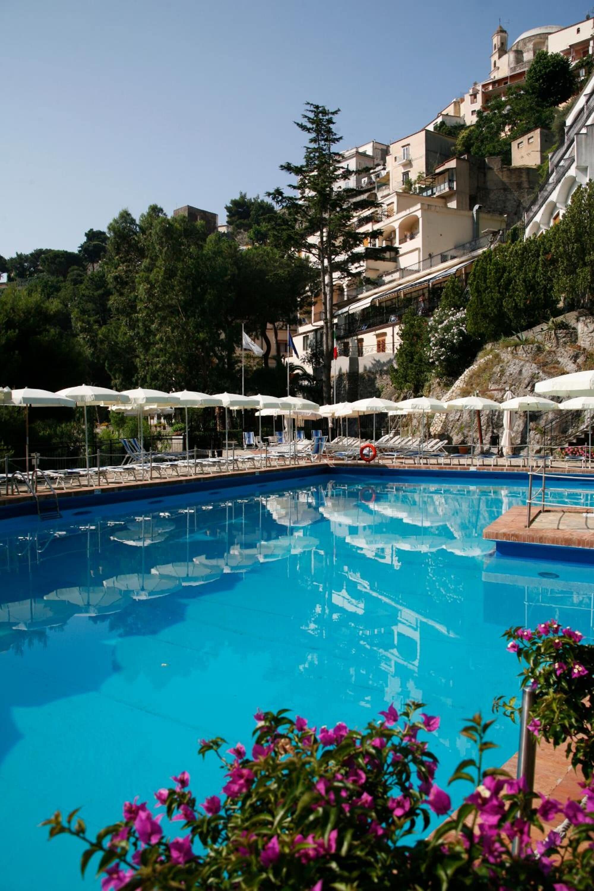 A swimming pool with sun loungers and umbrellas, surrounded by buildings and greenery.