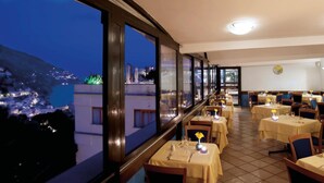 A restaurant with tables set for dining, large windows offering a view of a coastal town at night. - Hotel Royal Positano (Positano)