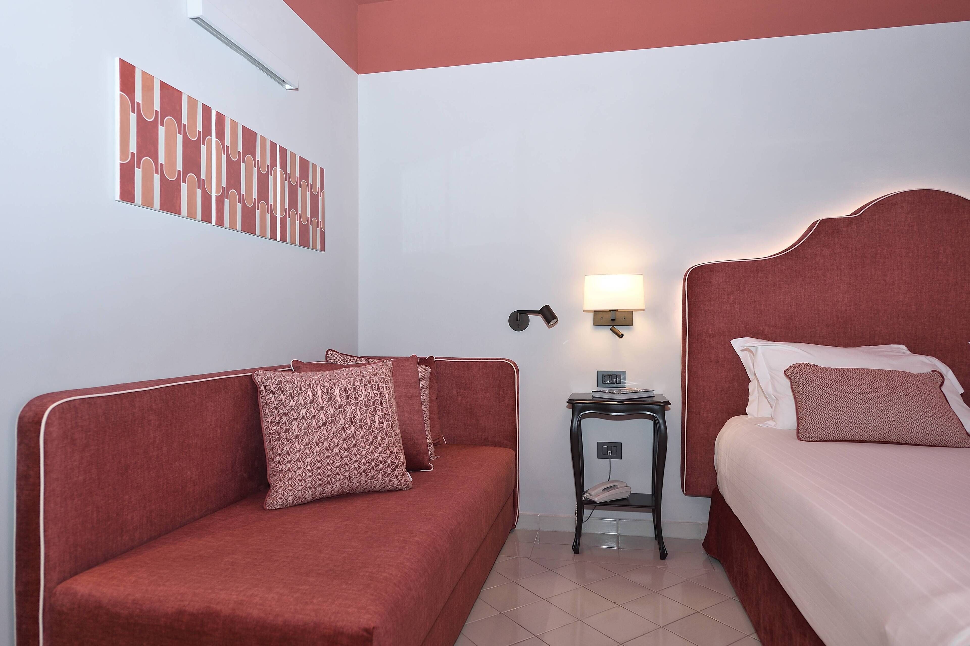 A room with a red sofa, a bed with a headboard, a bedside table with a lamp, and a wall art piece.