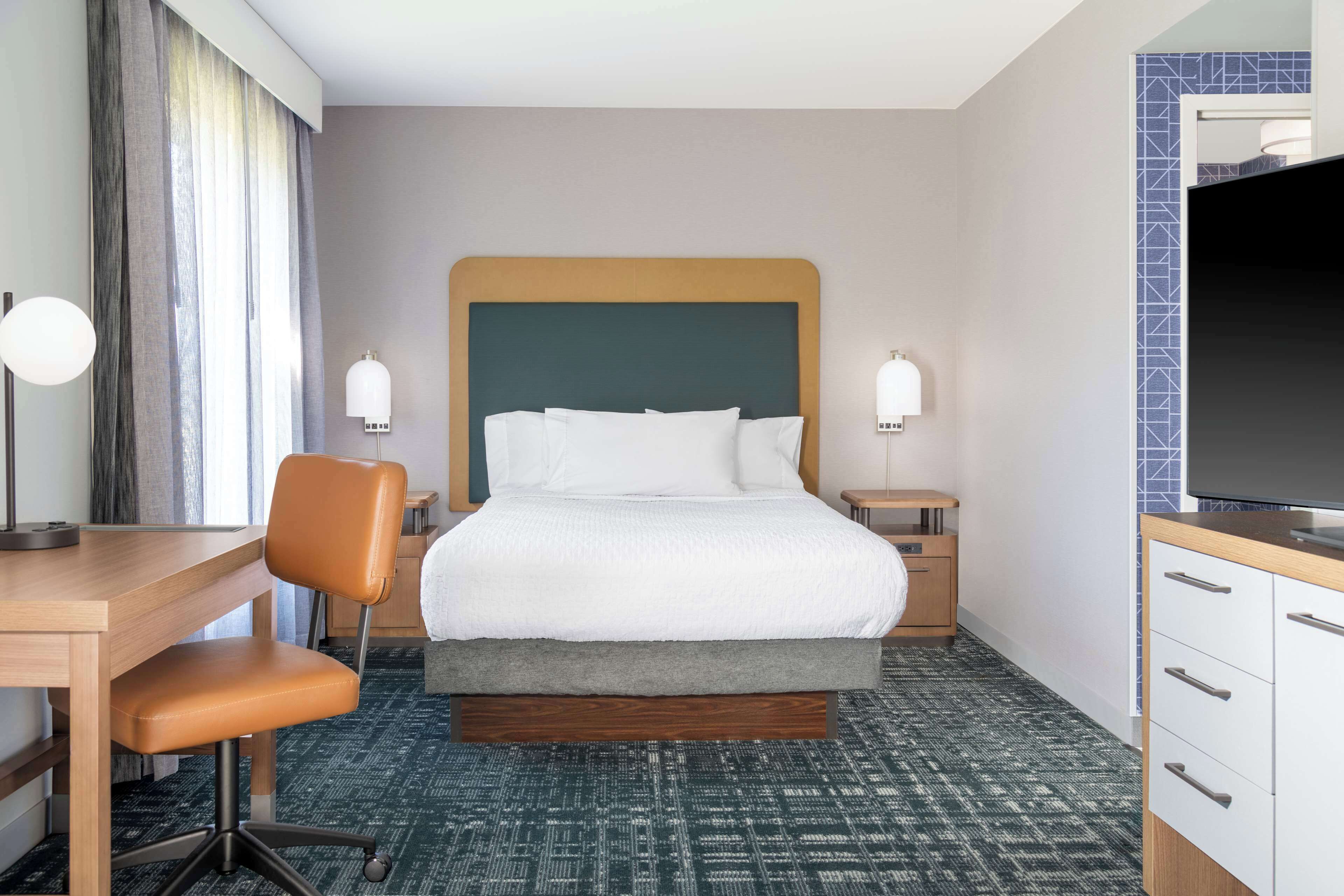 A hotel room with a bed, a desk, a chair, two bedside tables, a TV, and a patterned carpet.