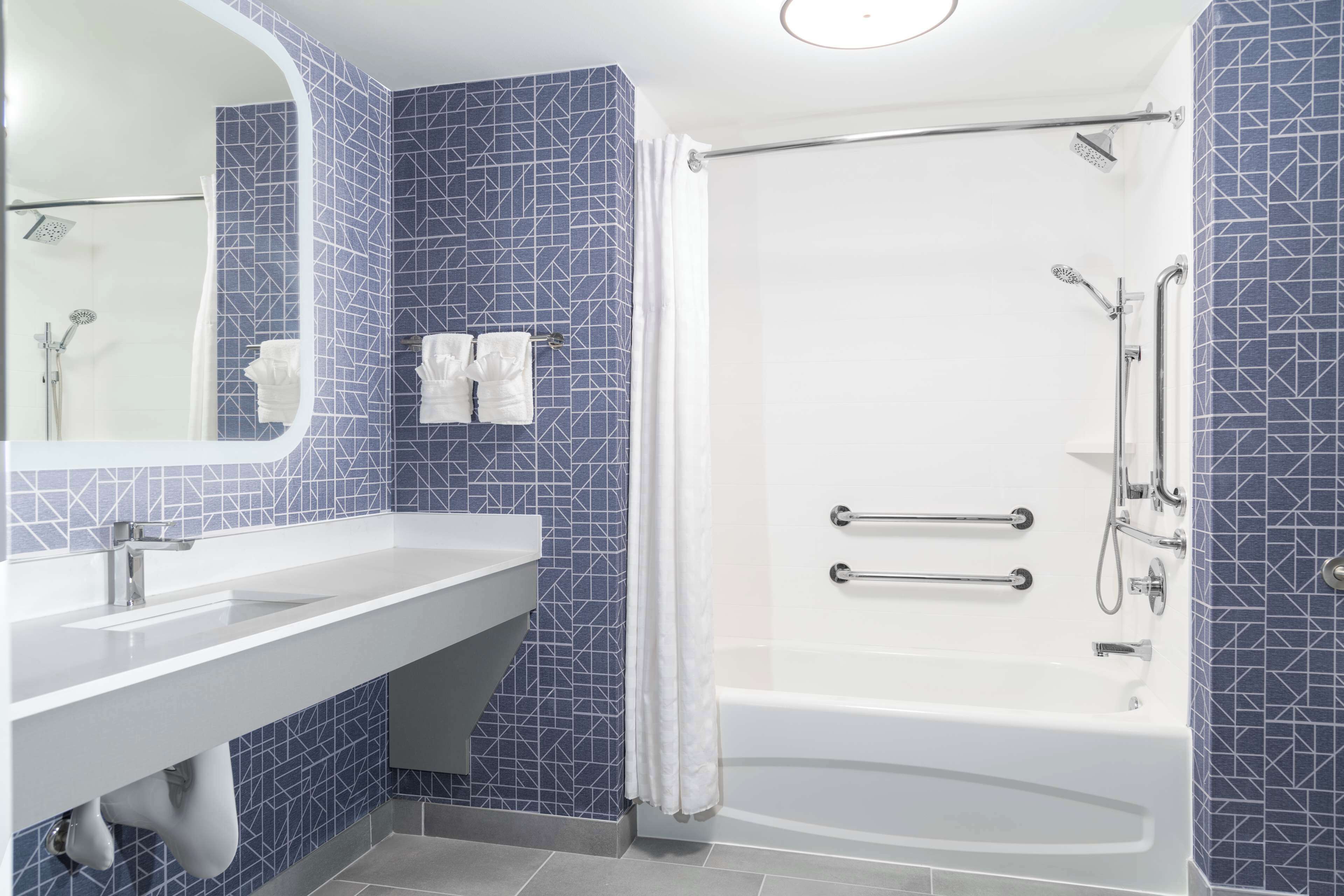 Un baño moderno con lavamanos blanco, bañera con barras de apoyo y ducha con cabezal de ducha de mano.