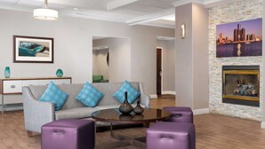 A modern living room with a grey sofa, blue cushions, a round coffee table, and purple ottomans. There is a fireplace with a cityscape painting above it. - Homewood Suites by Hilton Detroit-Troy (Troy)