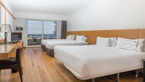 Habitación de hotel con dos camas, un escritorio, una silla y vistas al mar a través de amplios ventanales.