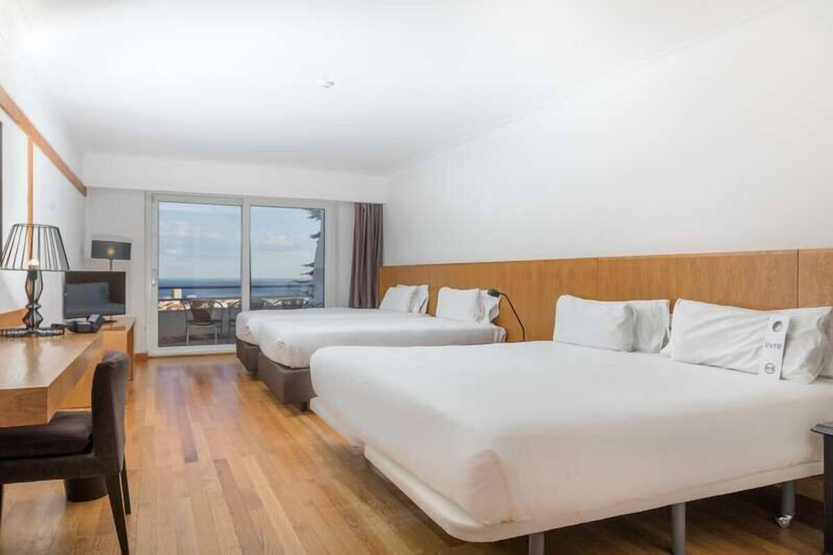 Une chambre d’hôtel avec deux lits, un bureau, une chaise et une vue sur la mer grâce à de grandes fenêtres.
