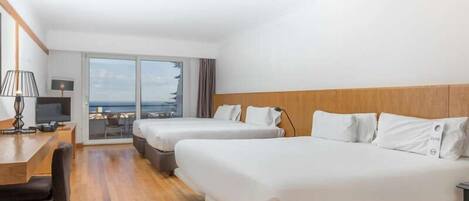 Habitación de hotel con dos camas, un escritorio, una silla y vistas al mar a través de amplios ventanales.