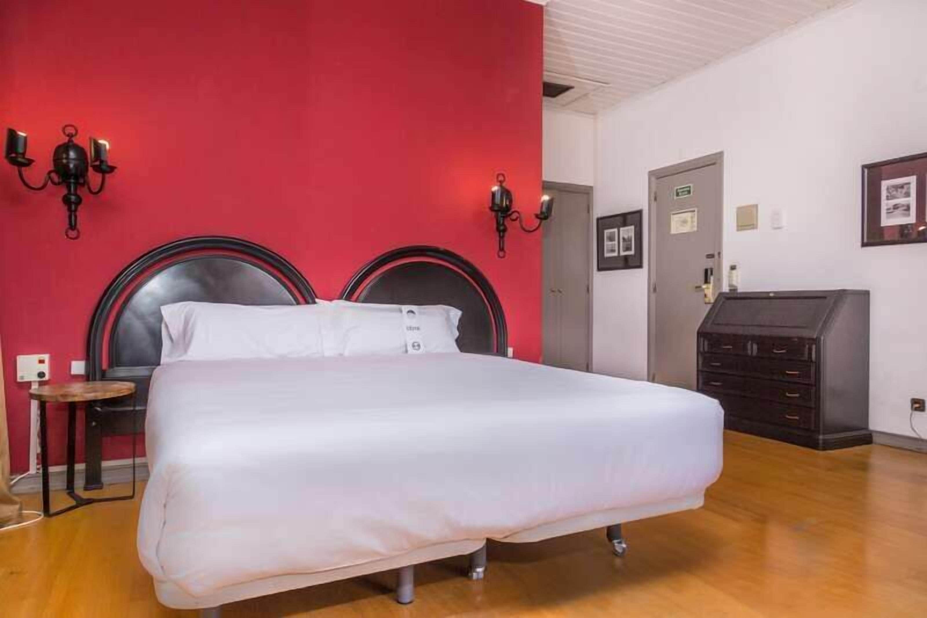 Une chambre d’hôtel avec un lit, une petite table, deux lampes fixées au mur, une commode et une porte.