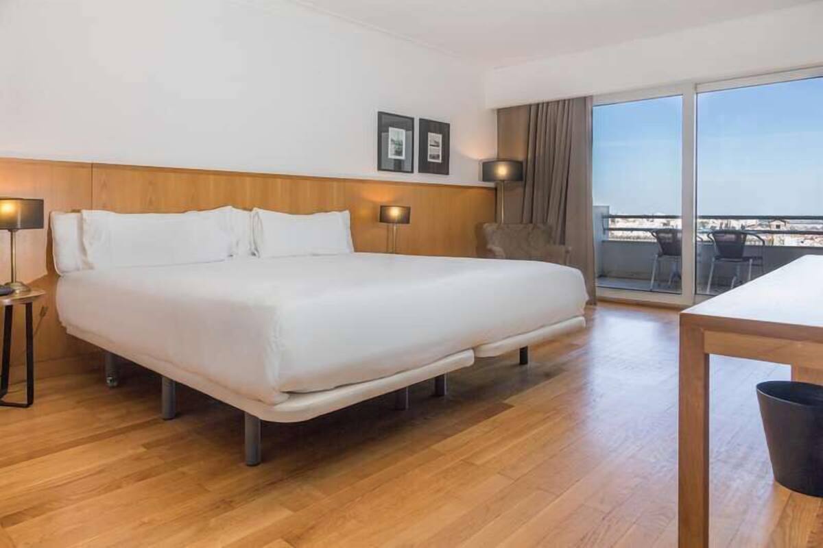 Une chambre d’hôtel moderne avec un grand lit, une tête de lit en bois, deux lampes de chevet, un fauteuil et un balcon avec vue.