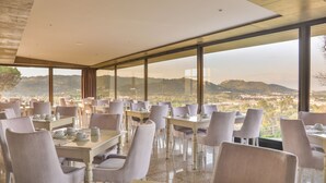 Un restaurante con mesas y sillas dispuestas para comer, ventanas amplias que ofrecen vistas a la ciudad y las colinas, y una decoración de estilo moderno.
