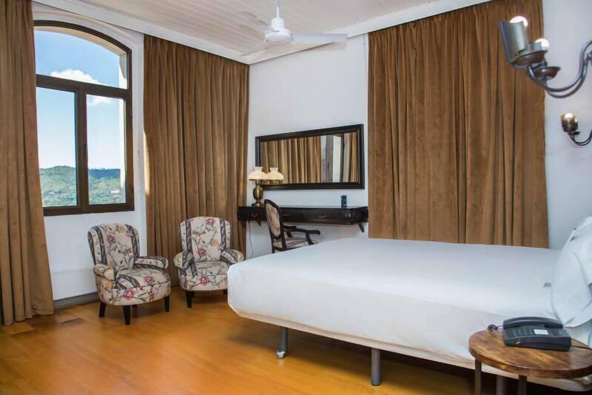 Une chambre d’hôtel avec un grand lit, deux fauteuils à motifs floraux, un bureau avec une lampe, un téléphone et une vue sur l’extérieur.