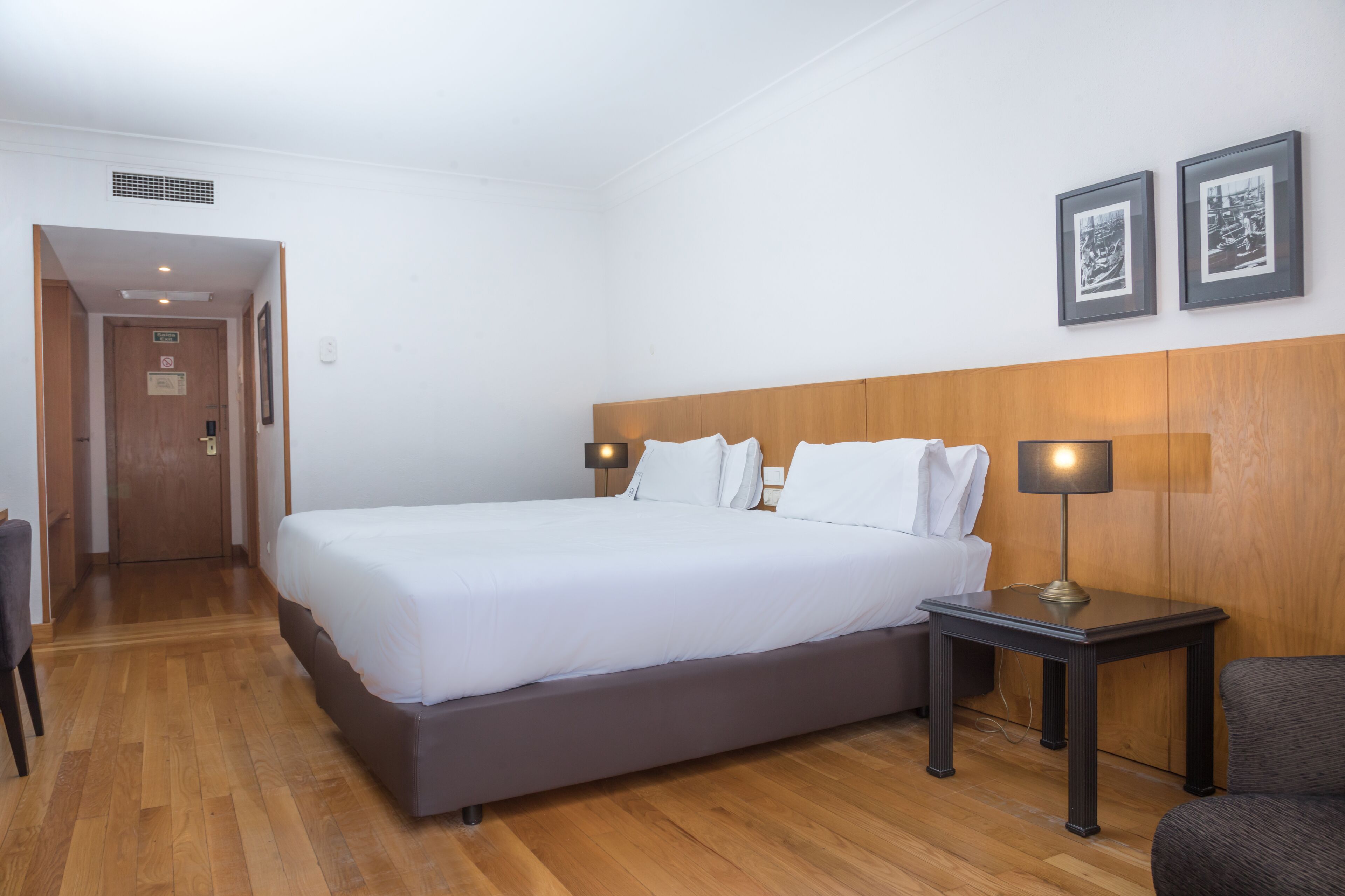 A hotel room with a large bed, two bedside tables with lamps, a wooden headboard, and two framed pictures on the wall.