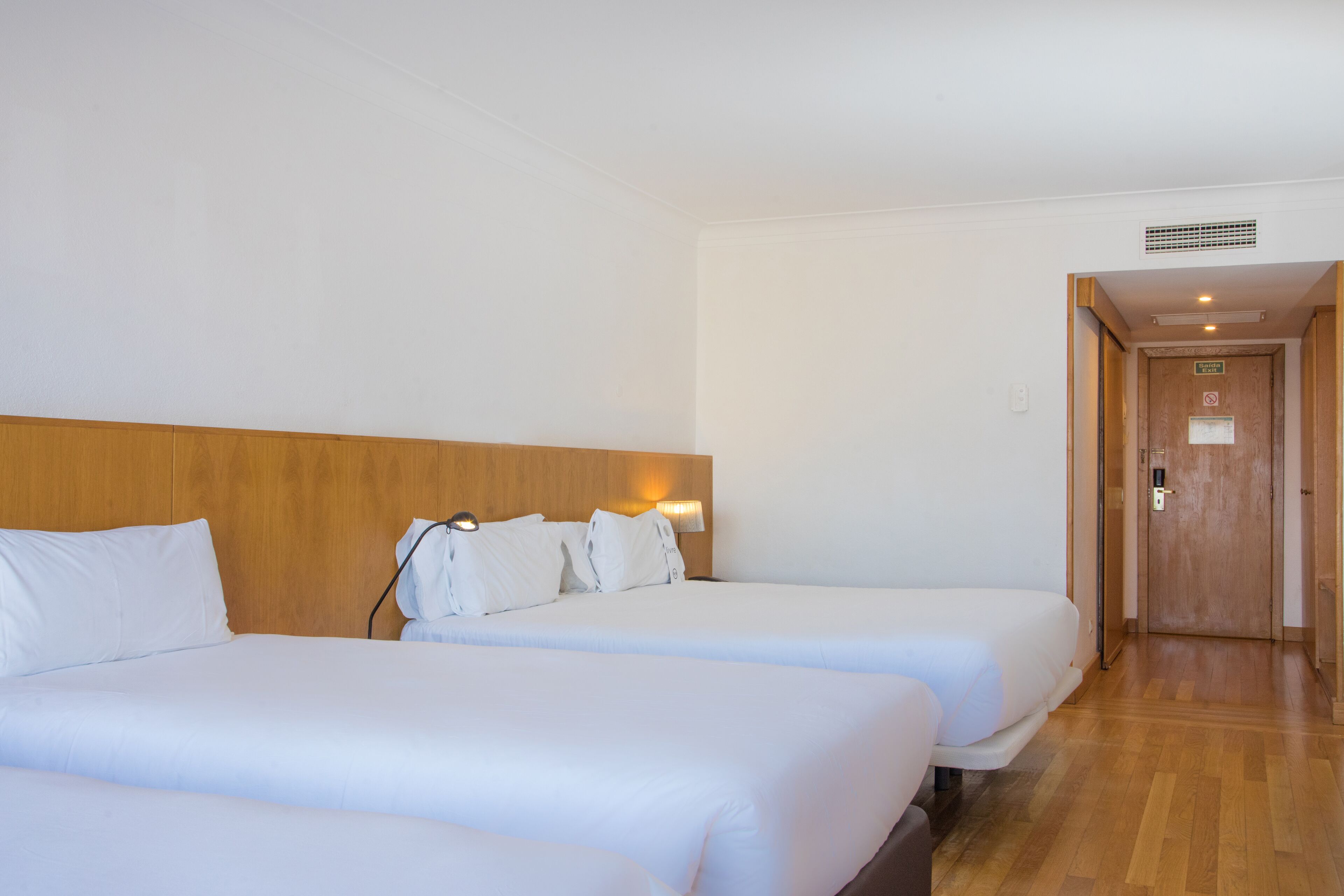 A hotel room with two beds, wooden headboard, white bedding, and a hallway leading to a door.