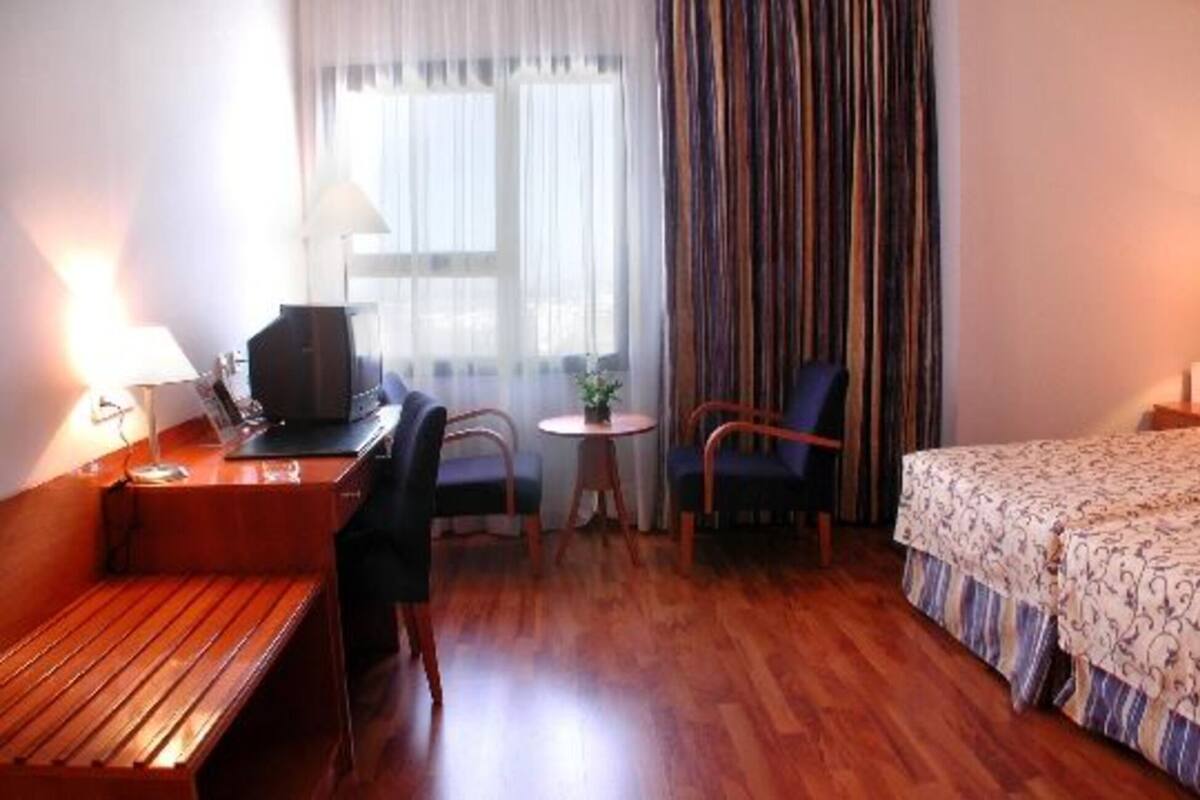Une chambre d’hôtel avec un lit, un bureau avec un ordinateur, une chaise, une petite table avec une plante et une fenêtre avec des rideaux.