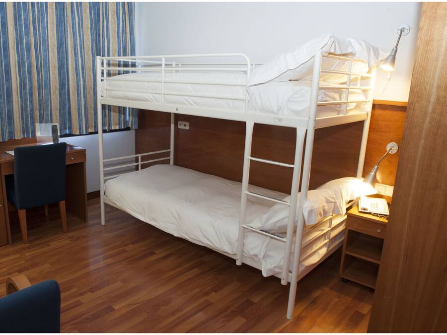 Uma cama beliche com escrivaninha e cadeira, um abajur e uma mesinha de cabeceira de madeira.