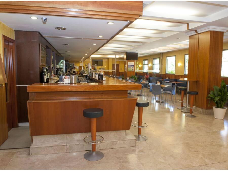 Amplio bar con mostrador de madera y dos taburetes, rodeado de diversas opciones de asientos y grandes ventanales.