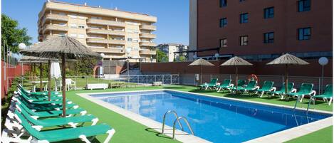 Una piscina con césped artificial verde, sillas de descanso y sombrillas.