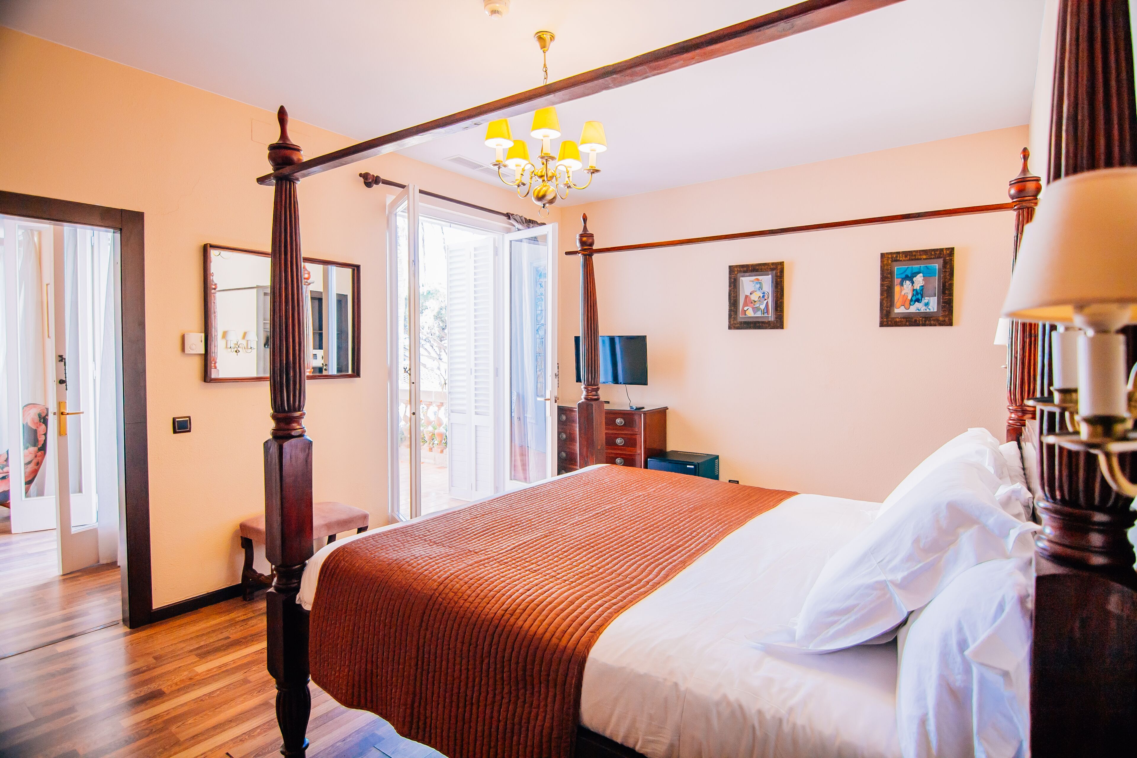 A four-poster bed with a wooden headboard, a chandelier, and a large window with blinds.