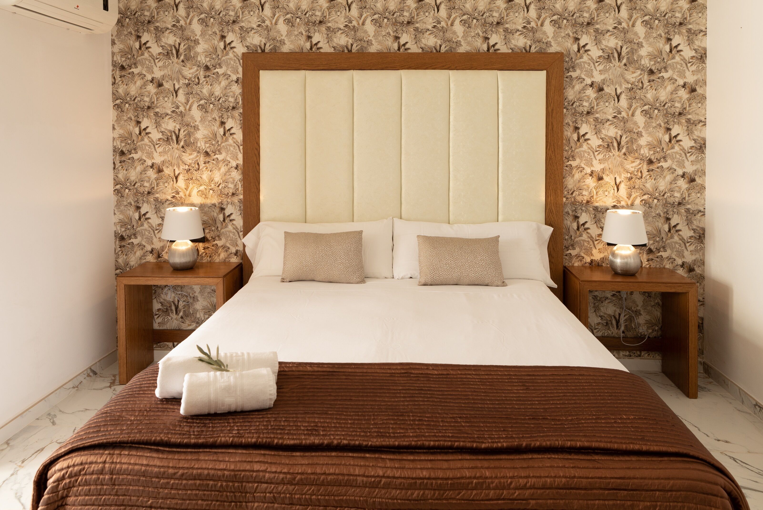 A neatly made bed with a brown quilt, two bedside lamps, and a floral wallpaper pattern.