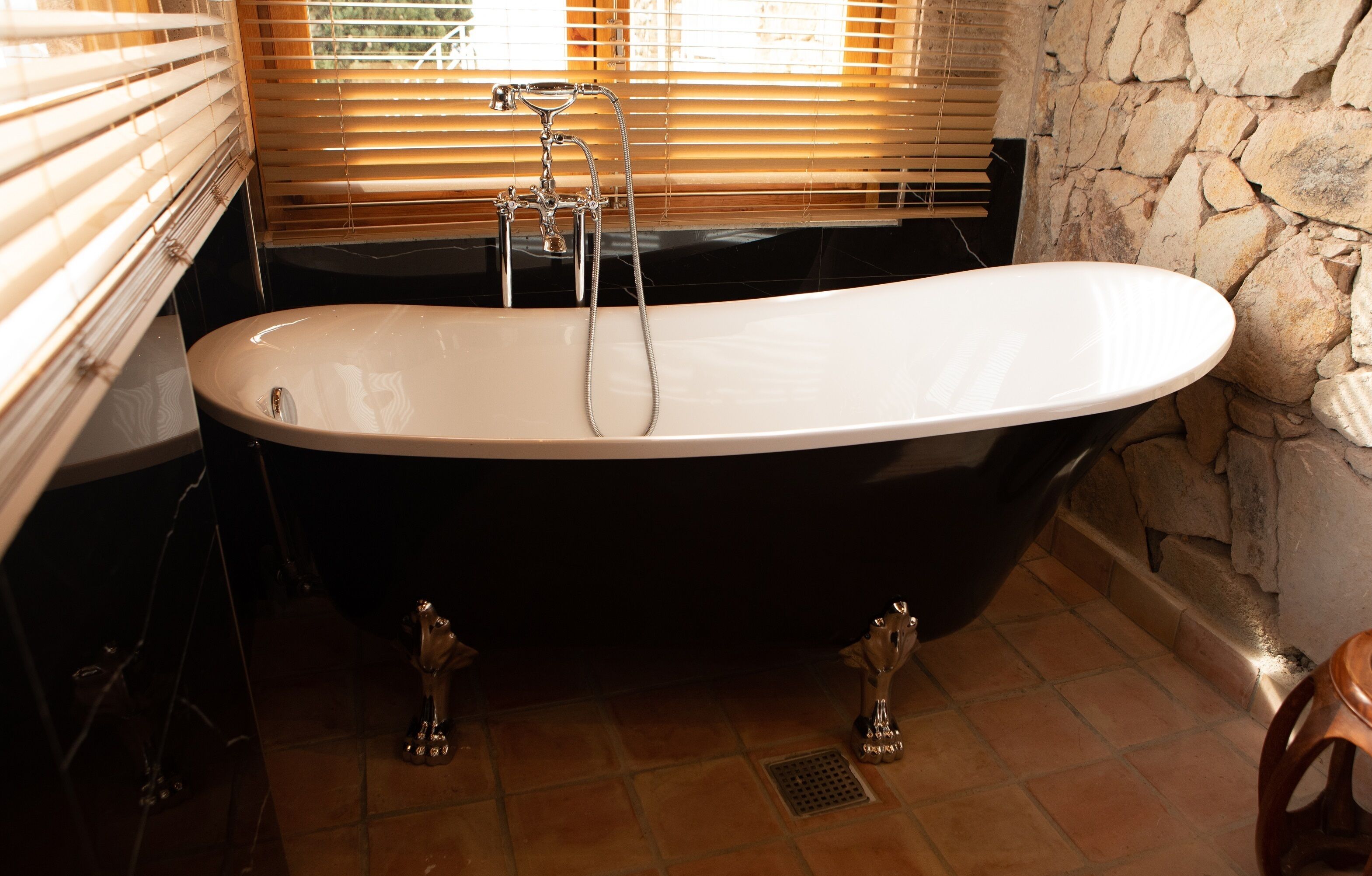 A black freestanding bathtub in a bathroom with a stone wall and a window with blinds.
