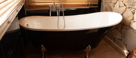 A black freestanding bathtub in a bathroom with a stone wall and a window with blinds.