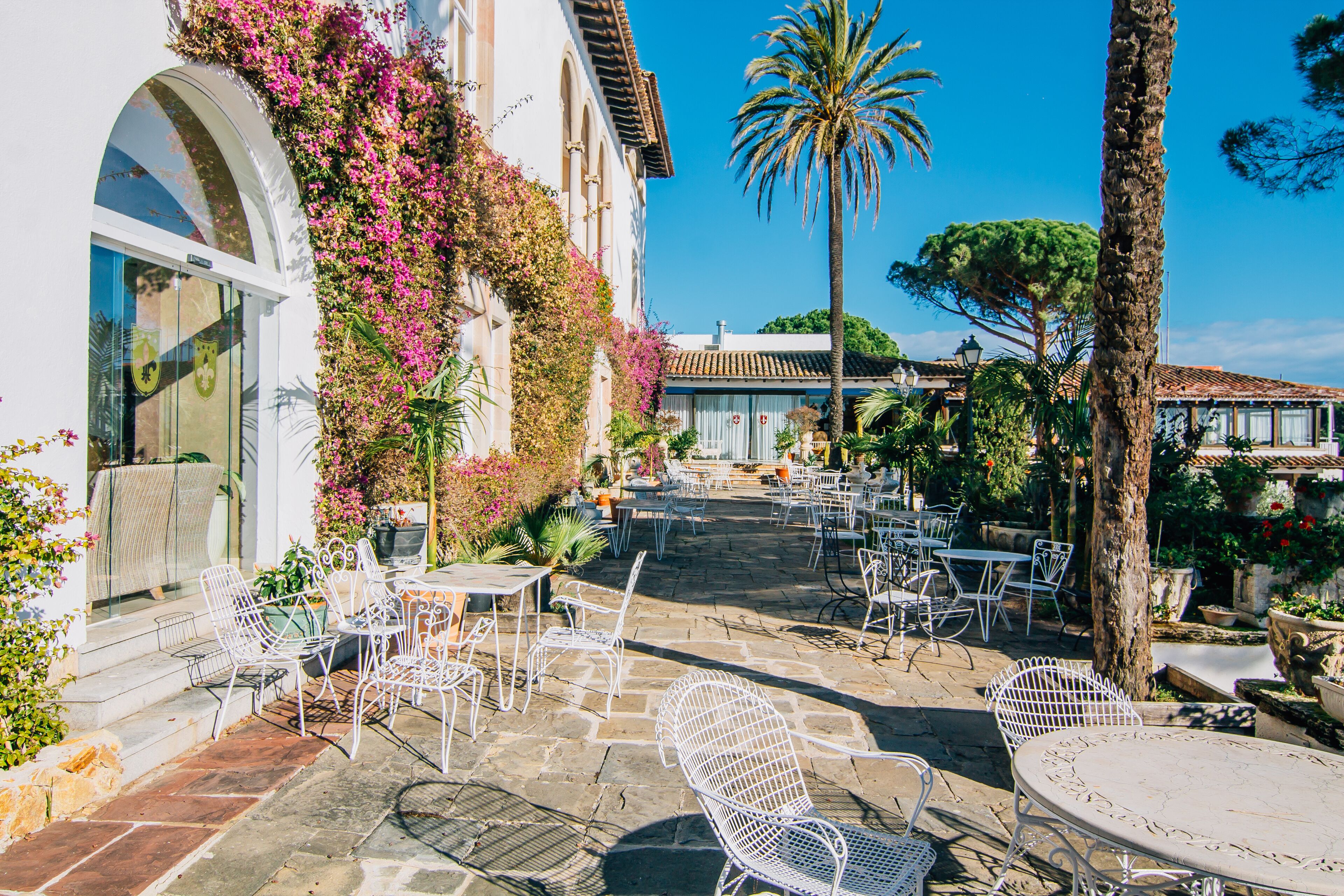 Une terrasse extérieure avec des chaises et des tables en métal blanc, entourée d’une végétation luxuriante et d’un palmier.