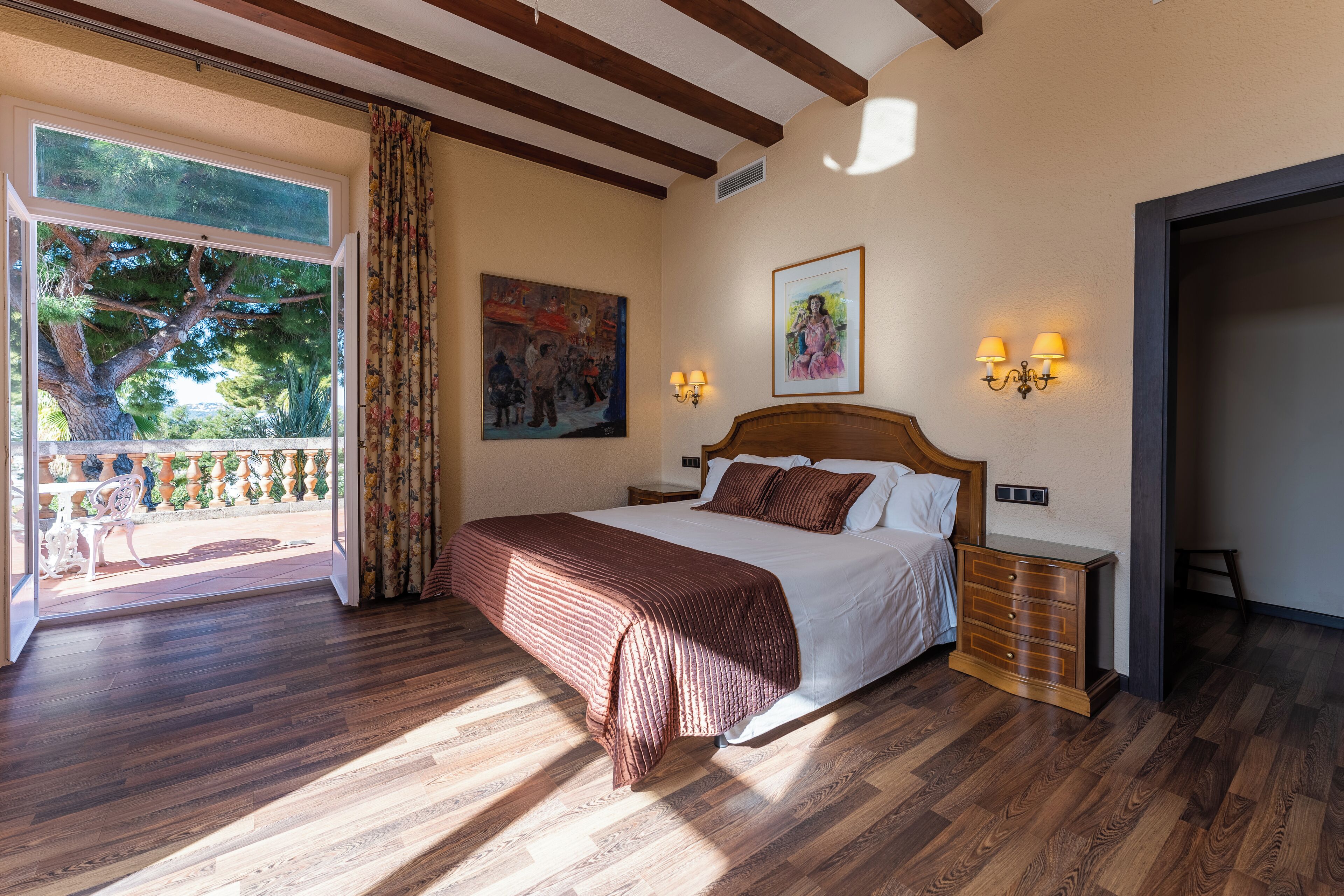 A bedroom with a large bed, wooden flooring, a balcony with a view of trees, and two wall-mounted lamps.
