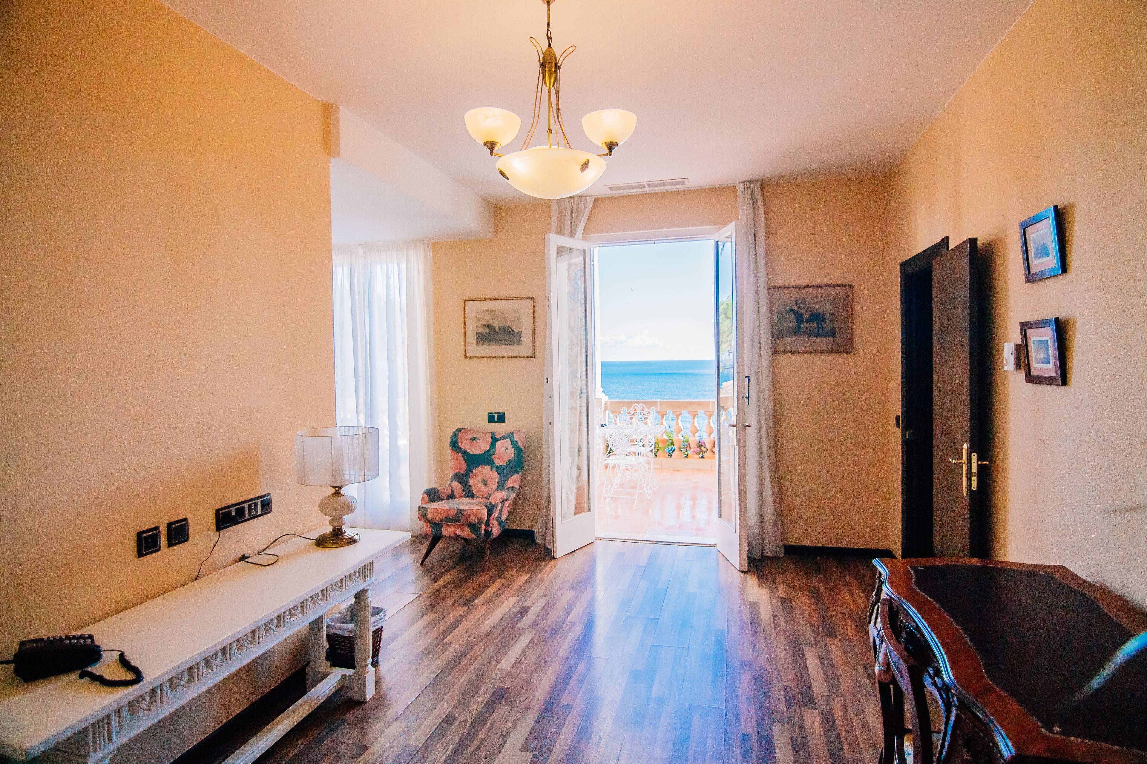 A room with a wooden floor, a white console table with a lamp, a chair with a floral pattern, and a door leading to a balcony with a view of the sea.