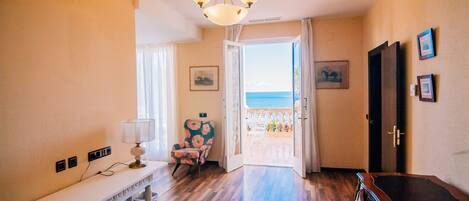 A room with a wooden floor, a white console table with a lamp, a chair with a floral pattern, and a door leading to a balcony with a view of the sea.