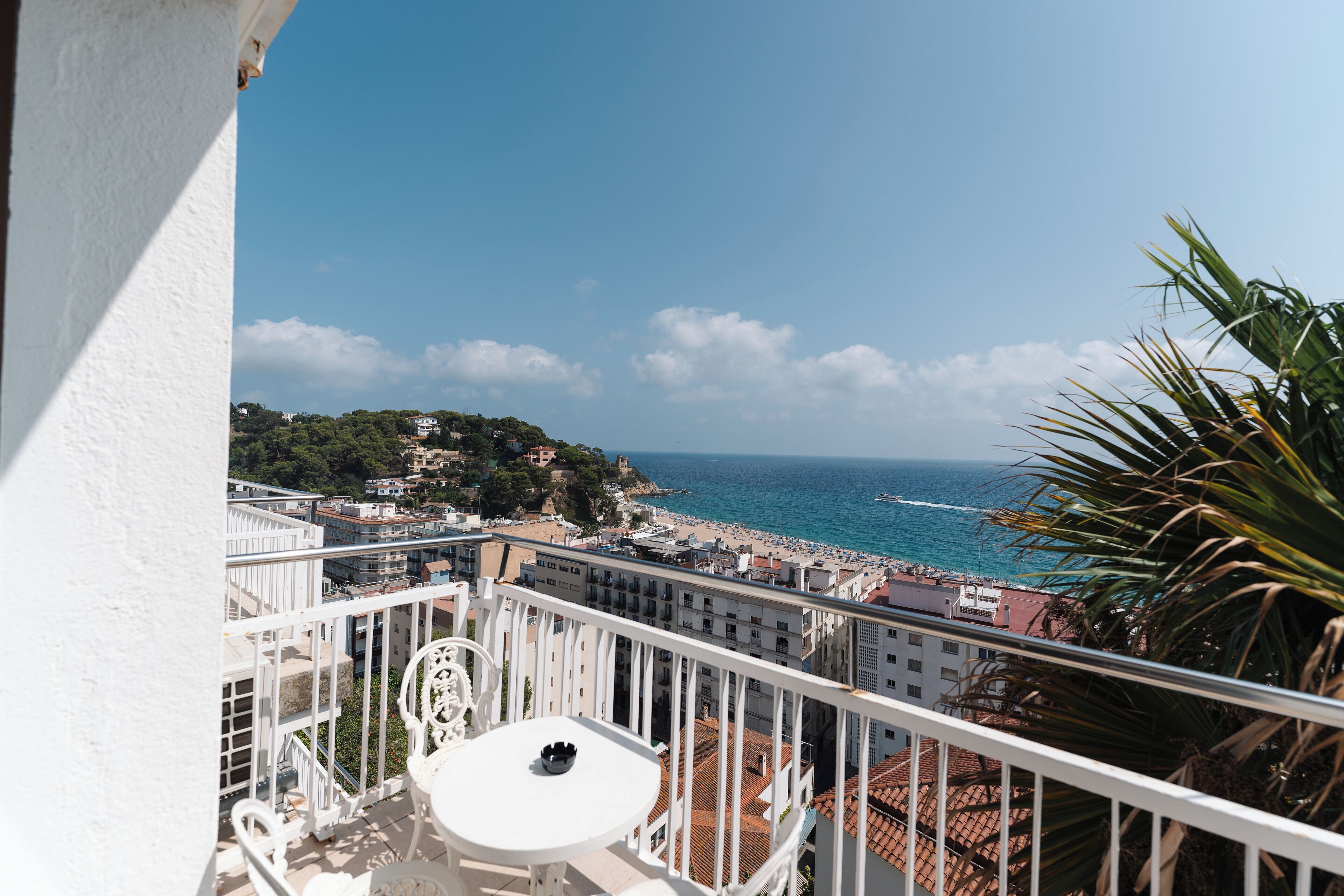 Varanda com mesa e cadeiras brancas, com vista para uma praia e uma vila costeira.