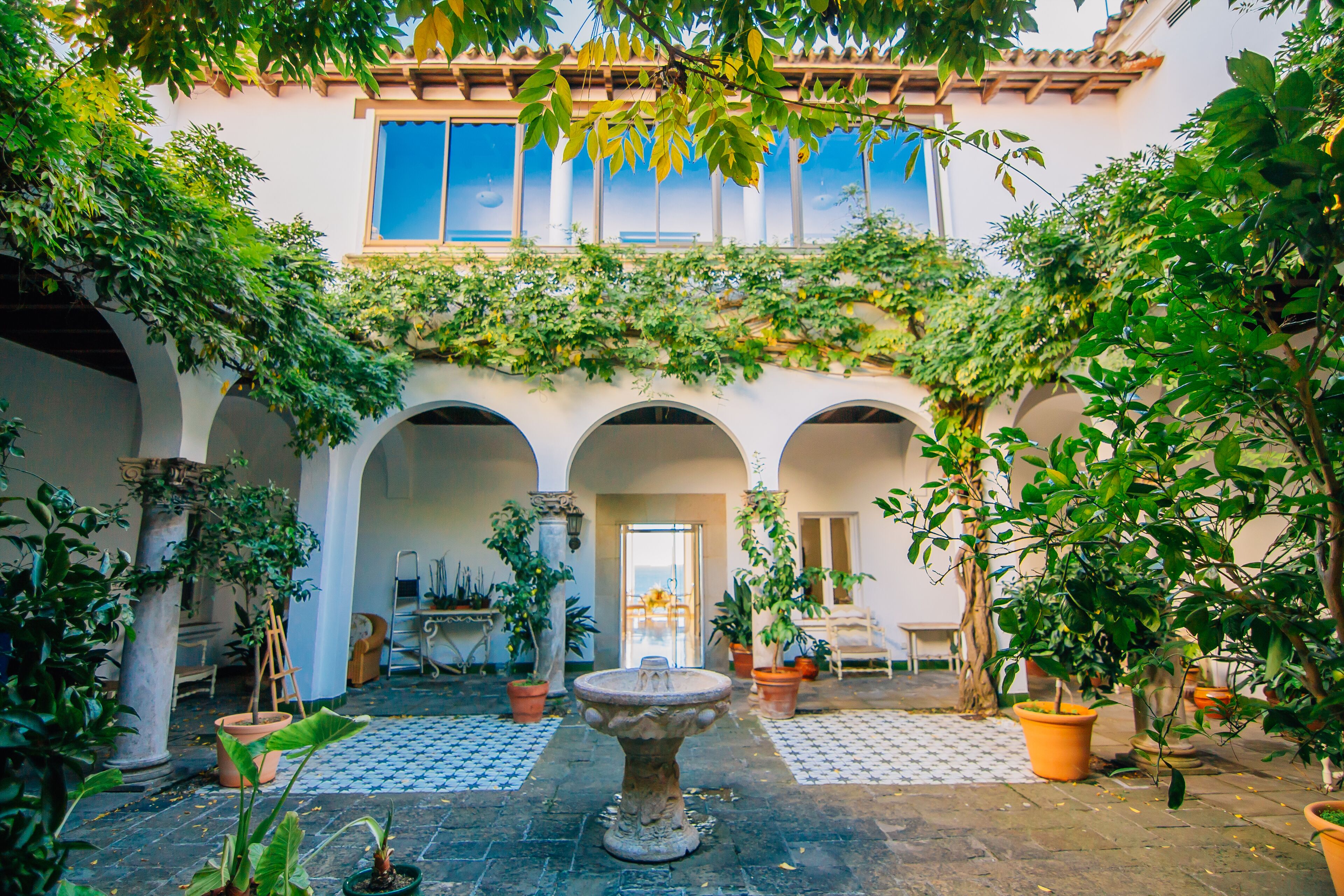 Un patio con arcos, plantas en macetas, una fuente de piedra y vistas de un edificio con ventanas amplias.
