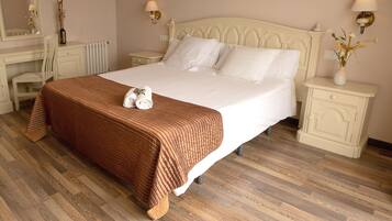 Una cama bien tendida con un edredón marrón, una almohada blanca y una toalla enrollada. Junto a la cama, una mesita de noche con una lámpara y un florero con flores secas. El cuarto tiene piso de madera y un tocador blanco con un espejo encima.