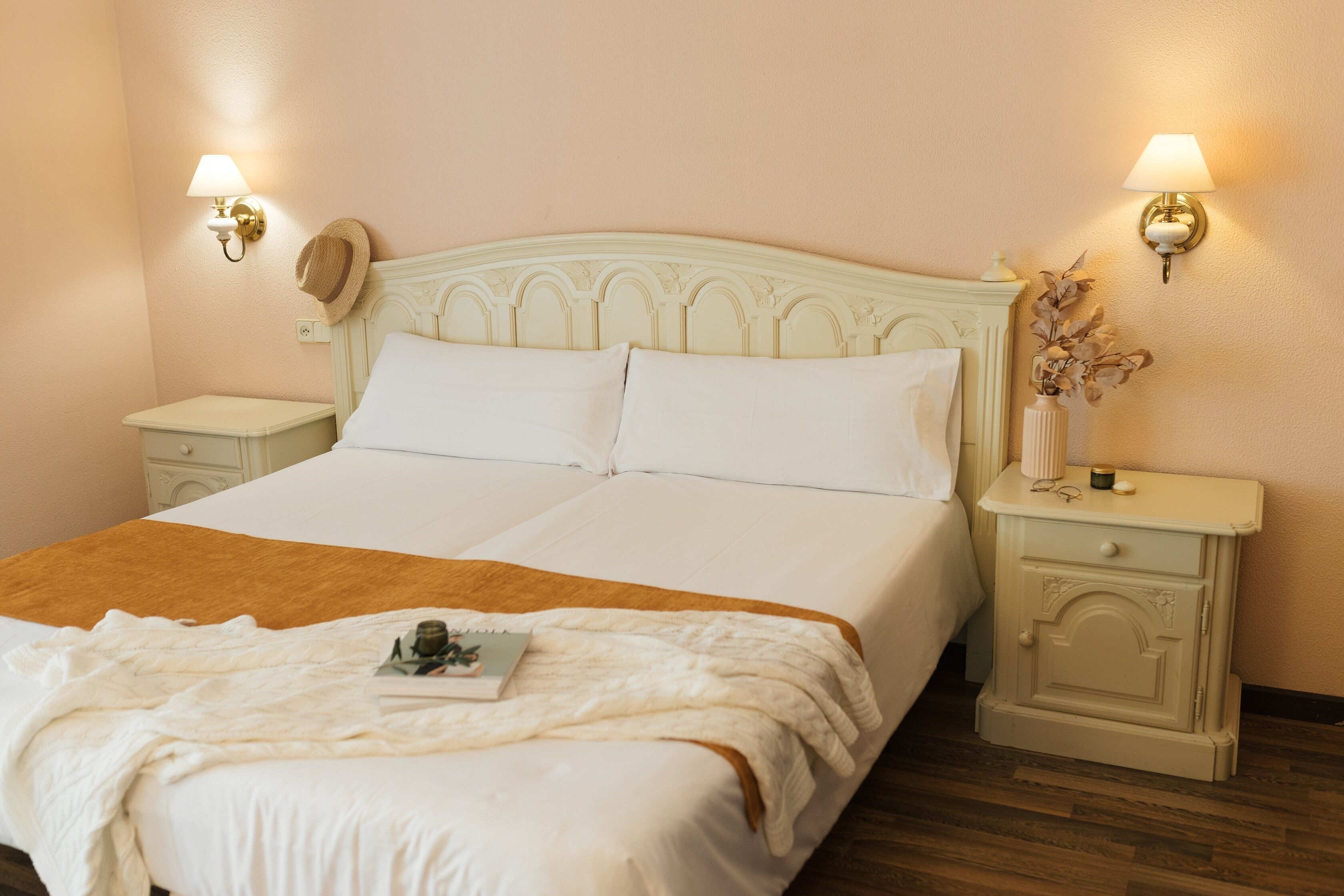 Una cama bien hecha con sábanas blancas y una manta marrón, dos mesitas de noche con lámparas, y un perchero de pared.