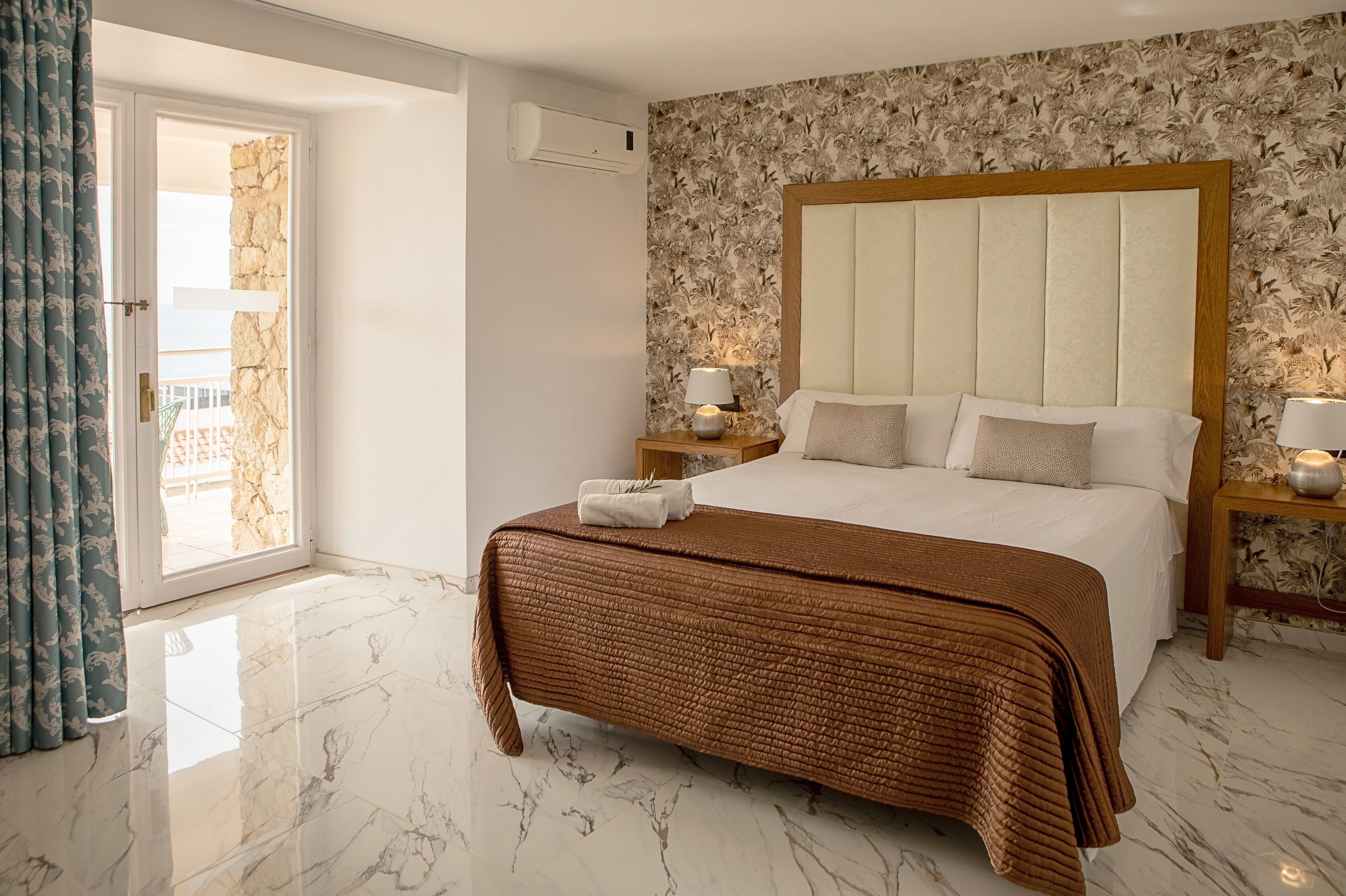 A bedroom with a large bed, two bedside tables with lamps, a floral wallpaper, and a view through a glass door.