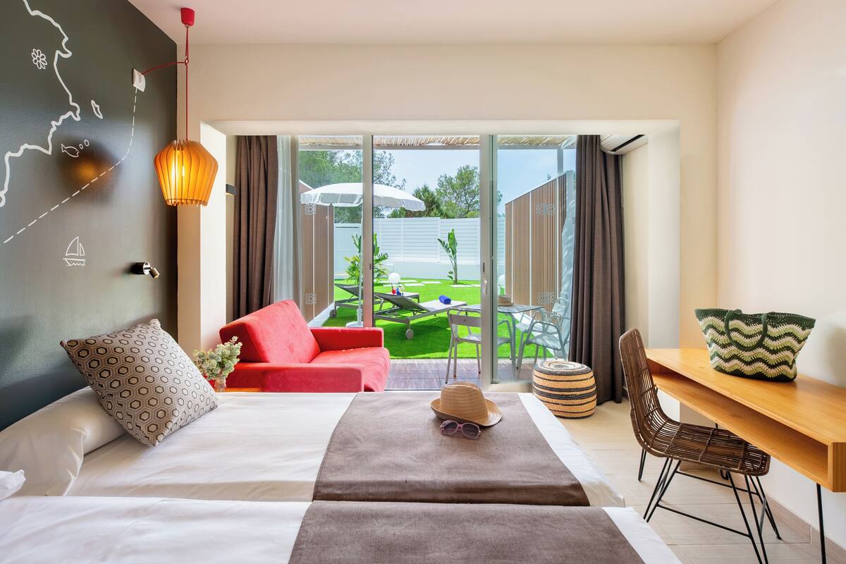 A modern hotel room with a bed, a red sofa, a dining table, and a view of a green garden through large sliding doors.