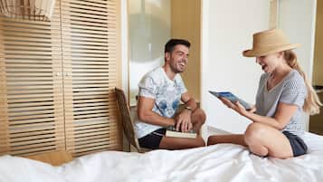 A couple sitting on a bed in a room with wooden blinds and a wicker chair.