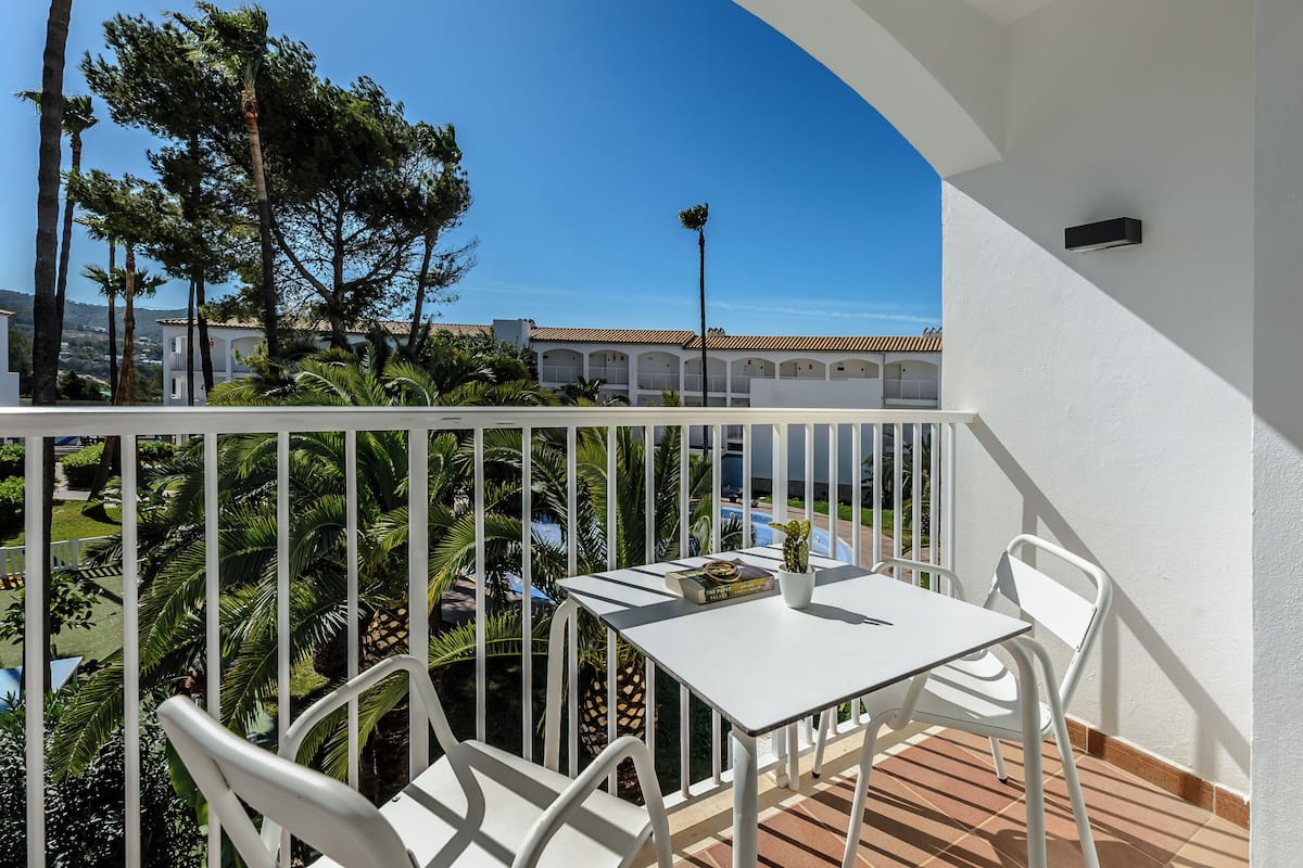 A balcony with a white table and chairs, overlooking a pool and a building with a tiled roof.