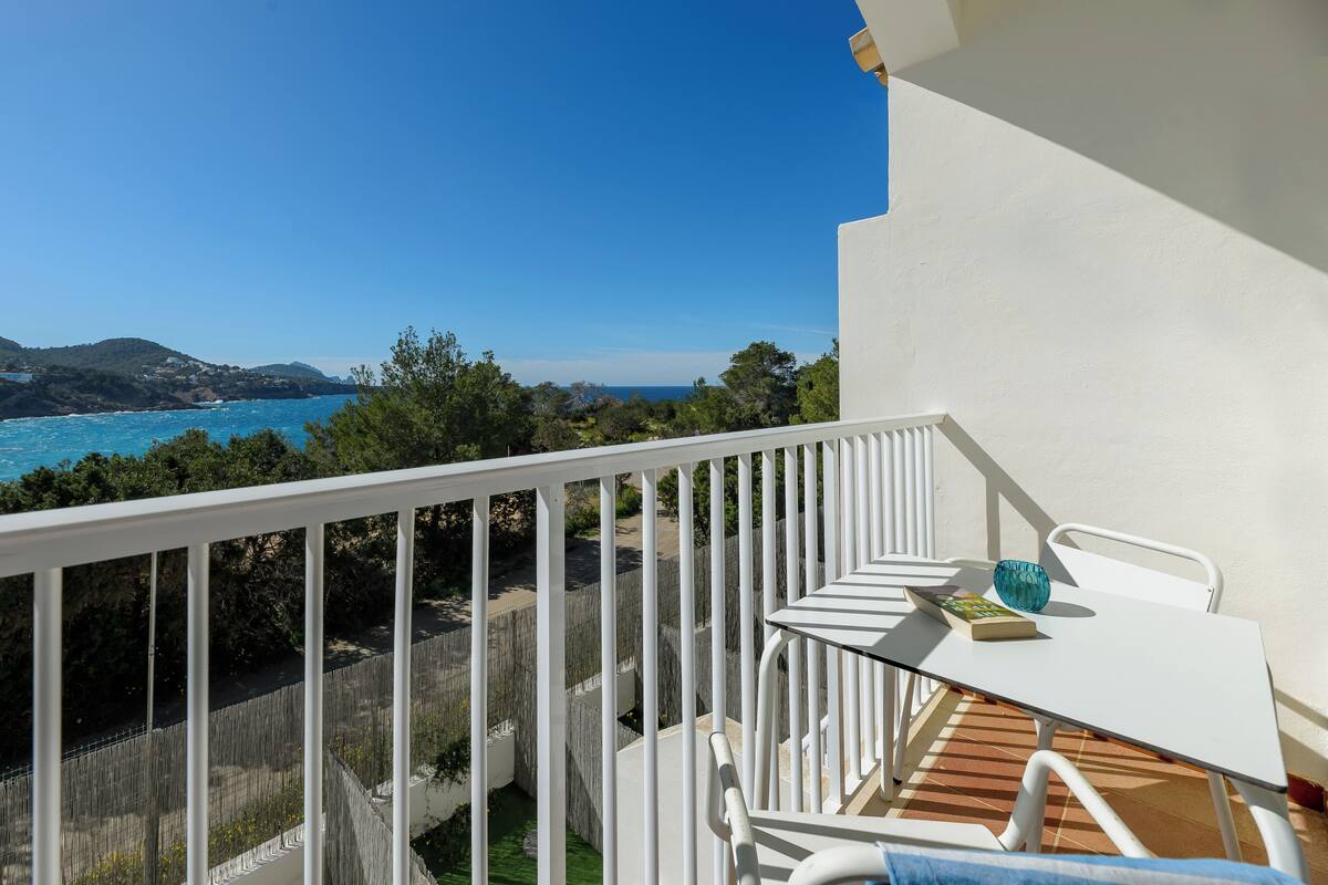 A balcony with a white railing, a small table with a book and a blue vase, and a view of the sea and hills.