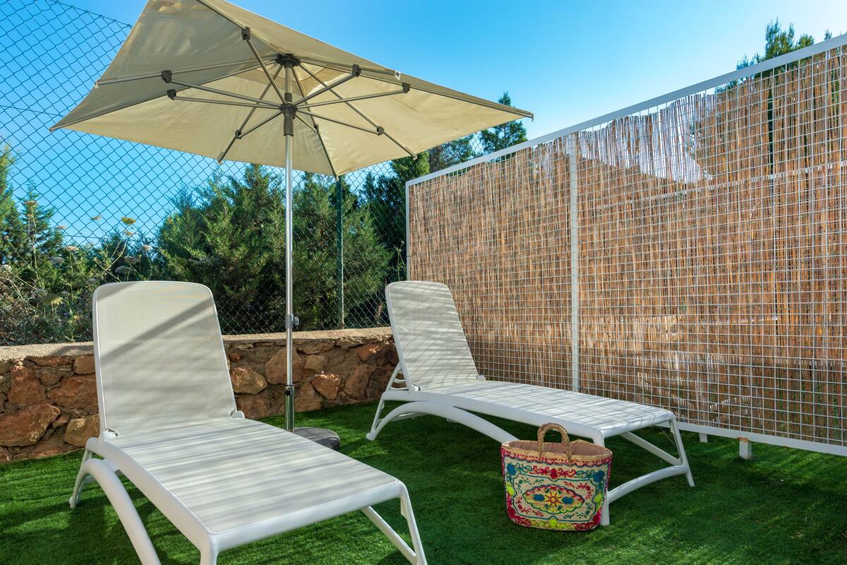 Two white lounge chairs with a beige umbrella and a colorful woven basket on a green lawn.