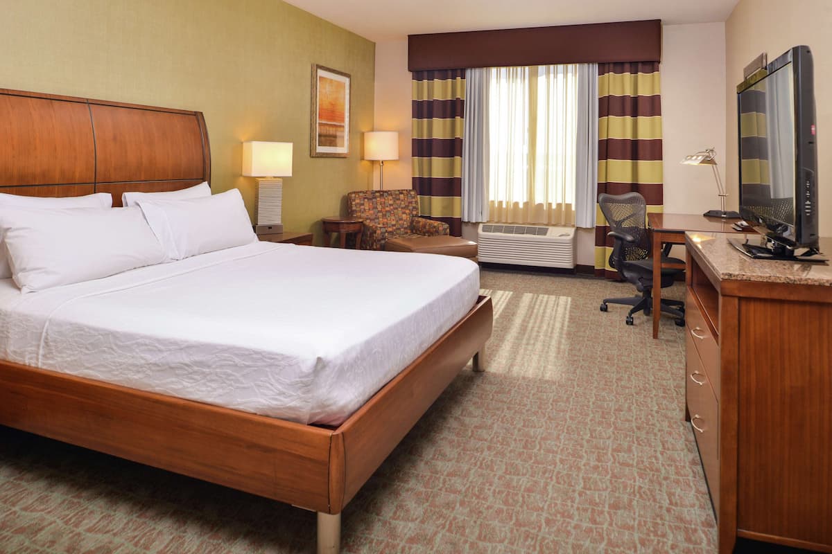 A hotel room with a large bed, a desk with a computer, a chair, an air conditioning unit, and a television.
