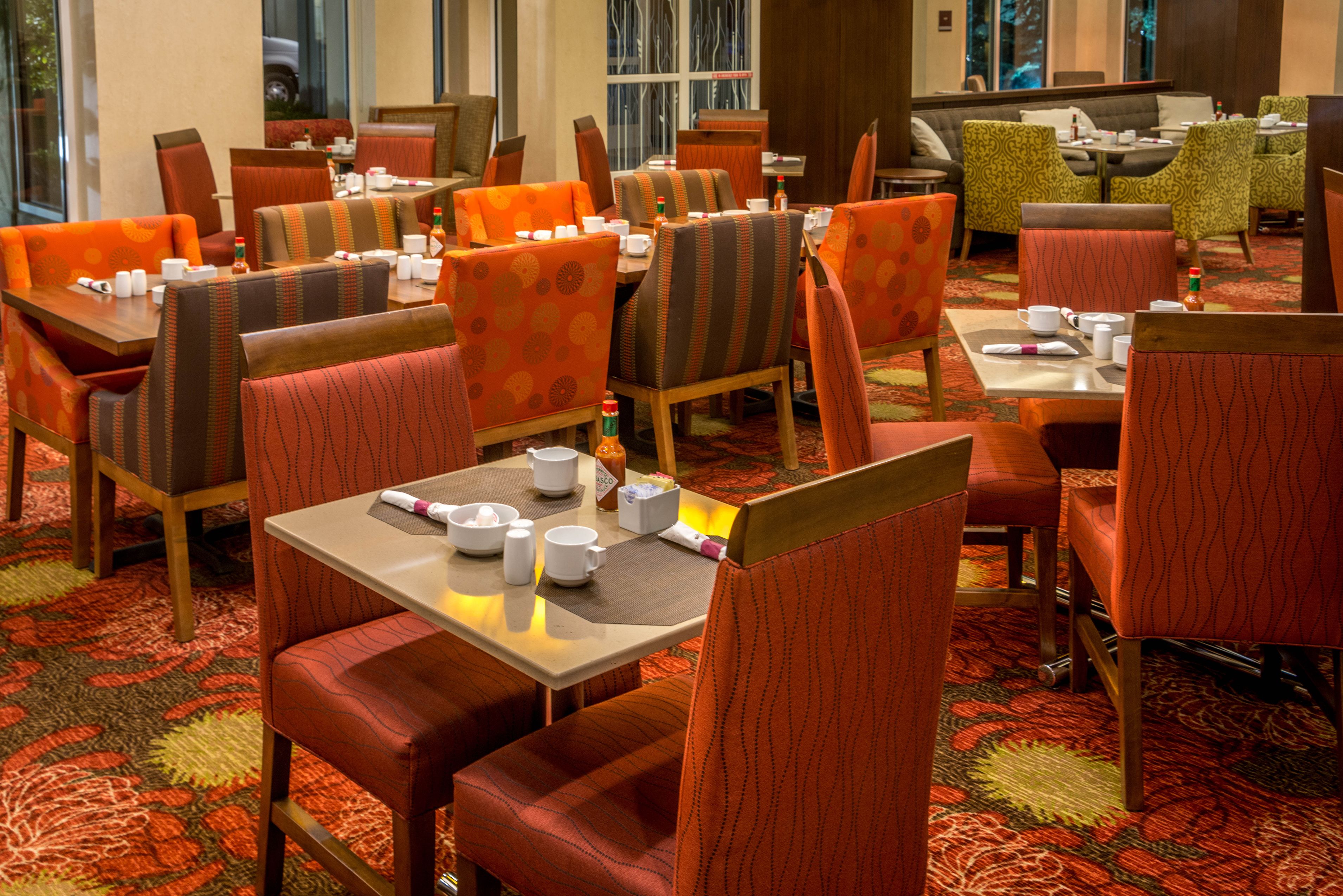 A hotel dining area with multiple tables and chairs, some with orange cushions.