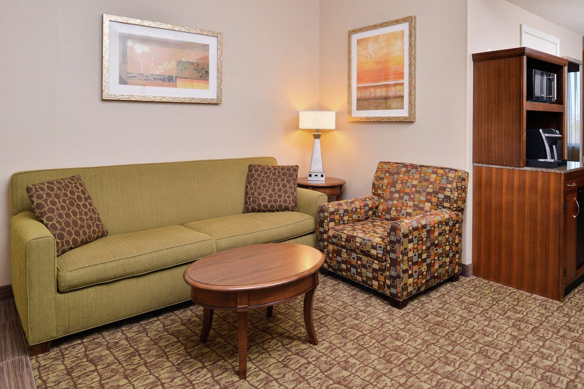 A living room with a green sofa, patterned armchair, wooden coffee table, and a lamp. There are framed pictures on the wall and a wooden cabinet with a microwave.