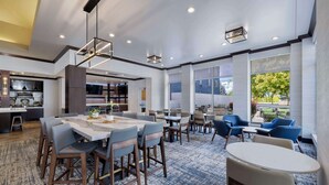A modern hotel lobby with a central dining area, a kitchen in the background, and large windows providing a view of the outdoors. - Hilton Garden Inn Boise Spectrum (Boise)