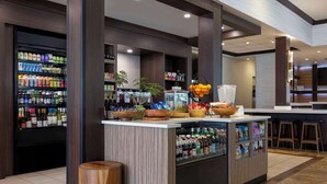 A well-lit bar area with shelves stocked with beverages and snacks, a central counter with a fruit bowl, and a seating area with stools. - Hilton Garden Inn Boise Spectrum (Boise)